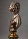Profile of a bronze bust of a woman, hair in twists swept back, gold earrings, beaded necklaces, and shoulders exposed in a dark brown wrapped top mounted on a gold base.