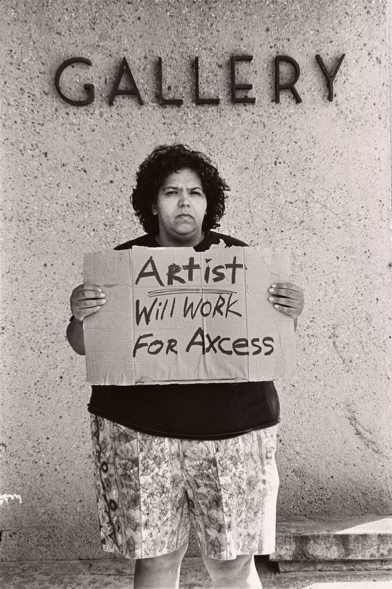 A grayscale photograph of a figure standing against a wall with the word “Gallery” above her head. Facing the camera, she holds a cardboard sign that reads “Artist will work for axcess,” spelled A-X-C-E-S-S.