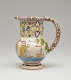 A jug with a rounded handle and pierced designs. The cream colored body is decorated with images of ships and a tree.
