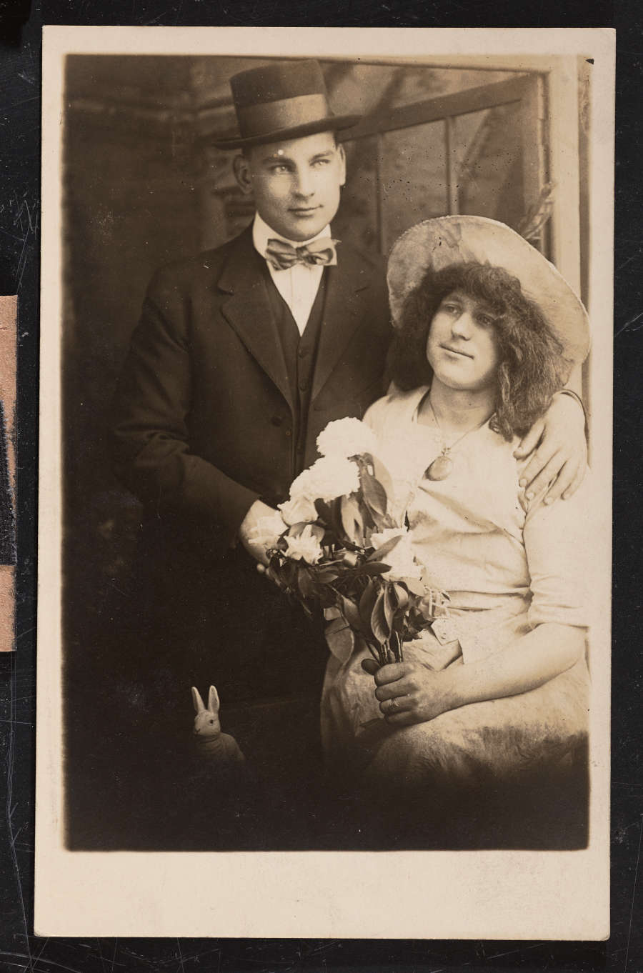 Photographic portrait of a man in a bowtie and hat. His arm is around another person, dressed as a woman, holding flowers. At bottom left, a small rabbit figurine appears. 