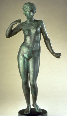 Gray metal sculpture with a green patina of a crowned nude woman standing on a circular pedestal, one arm down, grasping an object, and the other curled towards her shoulder. 