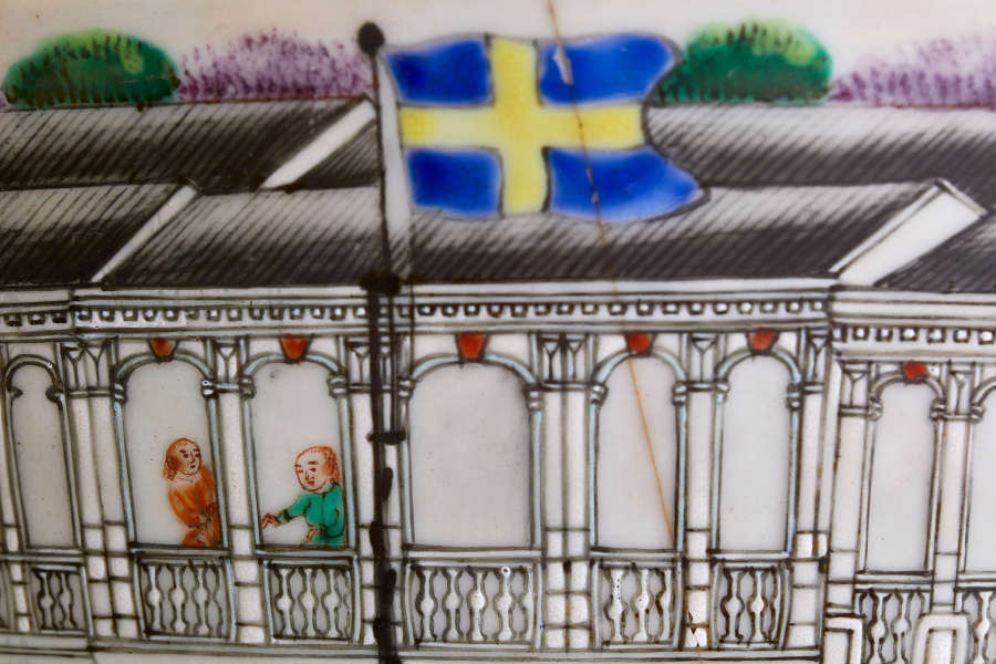 A close up of a painted ceramic showing figures along a building facade with arched windows and ornate barriers and a Swedish flag flying on a pole in the foreground.