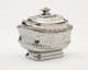 A silver sugar bowl with a decorative lid and a rounded square body with sculptural decorations.