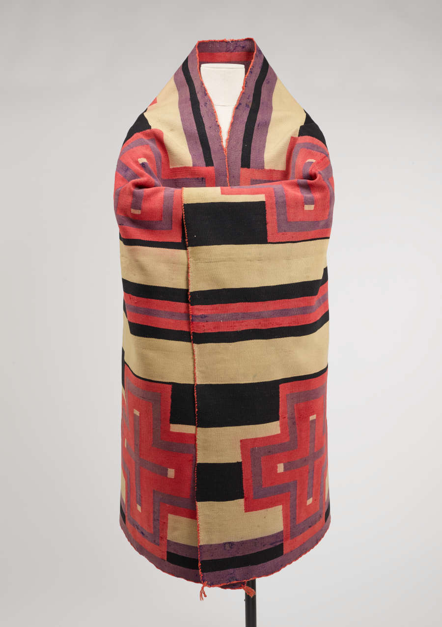 Striped woven blanket, worn as a robe, wrapped around a mannequin with folded arms. The stripes have varying widths and are different combinations of black, cream, purple, and pink.
