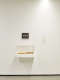  An installation photo depicting part of a white gallery room. A small black panel with illegible text is off-center on the wall, below, an enclosed shelf holds two rectangular objects.