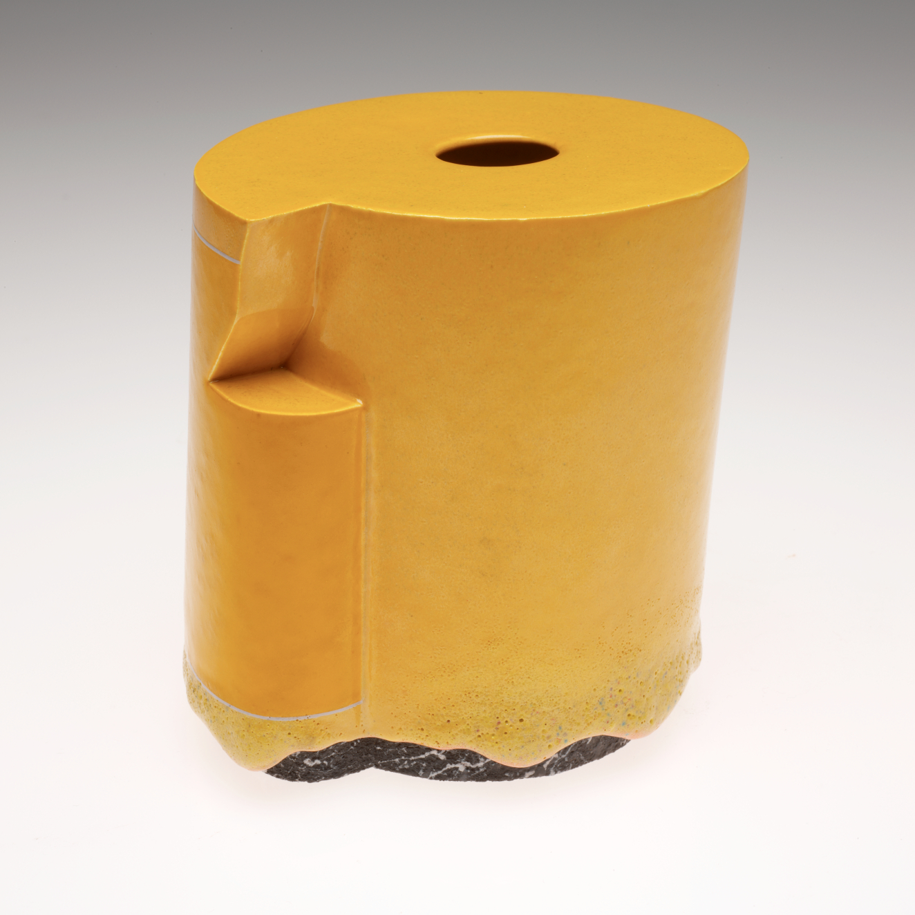 A glossy yellow cylindrical ceramic vessel with a small hole cut into the top, an irregular geometric cutout on the side, and a textured black base.