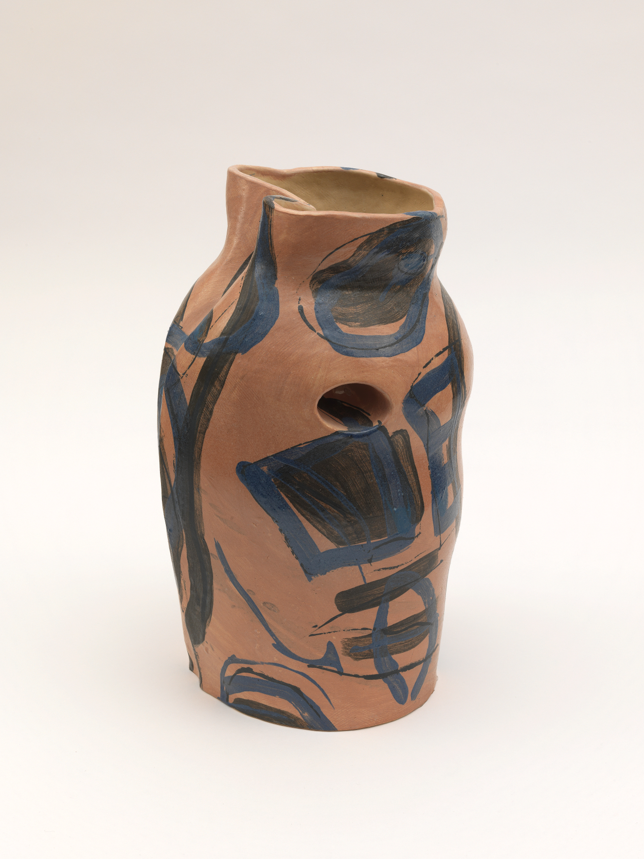 Asymmetrical clay vessel in warm pink tones with a small opening on its side, its uneven rim and subtly lopsided form painted with abstract blue and black geometric designs.