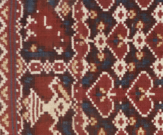 A detail of a textile with small white squares surrounding deep red diamonds in dense, organized patterns on a dark blue and red background. 