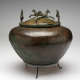 A brown-green patinated metal bowl with decorative engravings on its onion-shaped body, sitting on a three-legged stand. Metal-cast winged horses dance along the rim of the bowl’s domed lid.