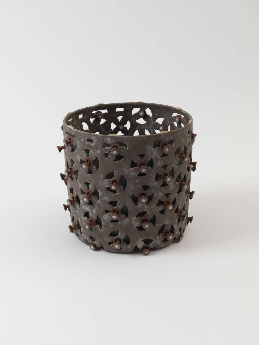 Dark brown cylindrical vessel with evenly spaced repeating triangular cutouts that form irregular circles. The center of each circle is punctuated by a raised round stud.