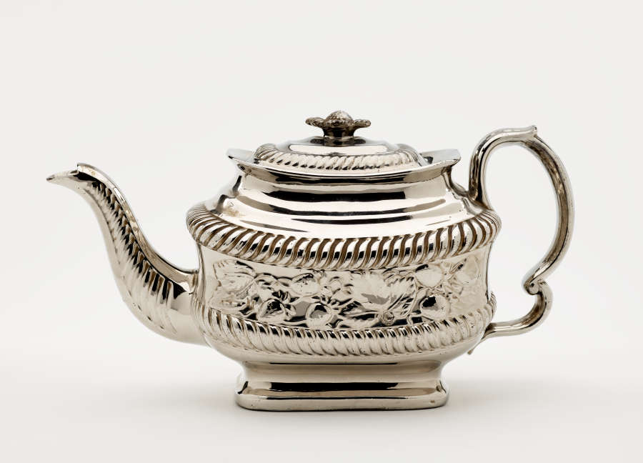 A silver teapot with a decorative handle, a rounded square body and spout with sculptural decorations.