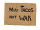 A horizontal, rectangular piece of creased cardboard that features centered black hand-written text reading: “Make Tacos Not War”.