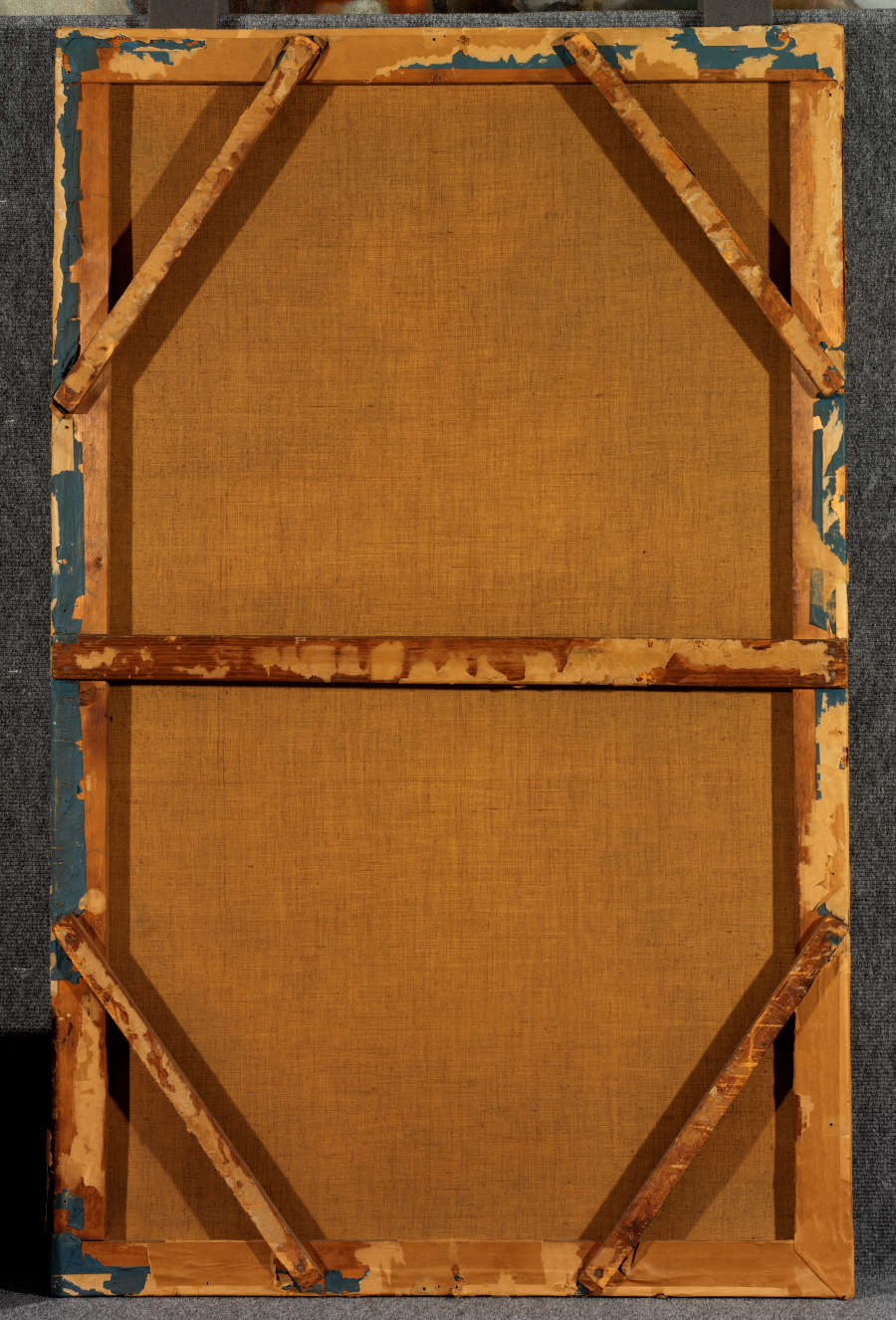 Back of yellowed stretched canvas with wooden frame, supported by diagonal braces at each corner, with chipped blue paint around the worn edges