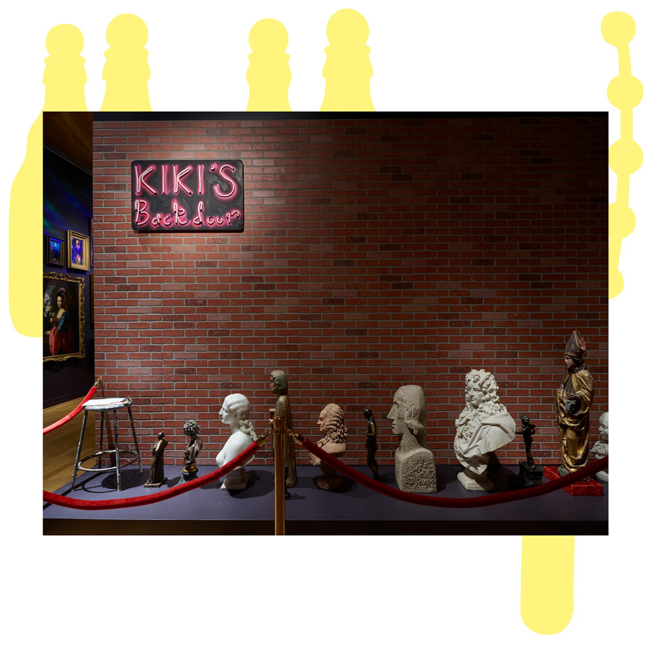 A brick wall bears a sign saying “Kiki’s Back Door.” Waiting on the ground behind a red velvet rope is a row of portrait busts and historical figurative artworks.