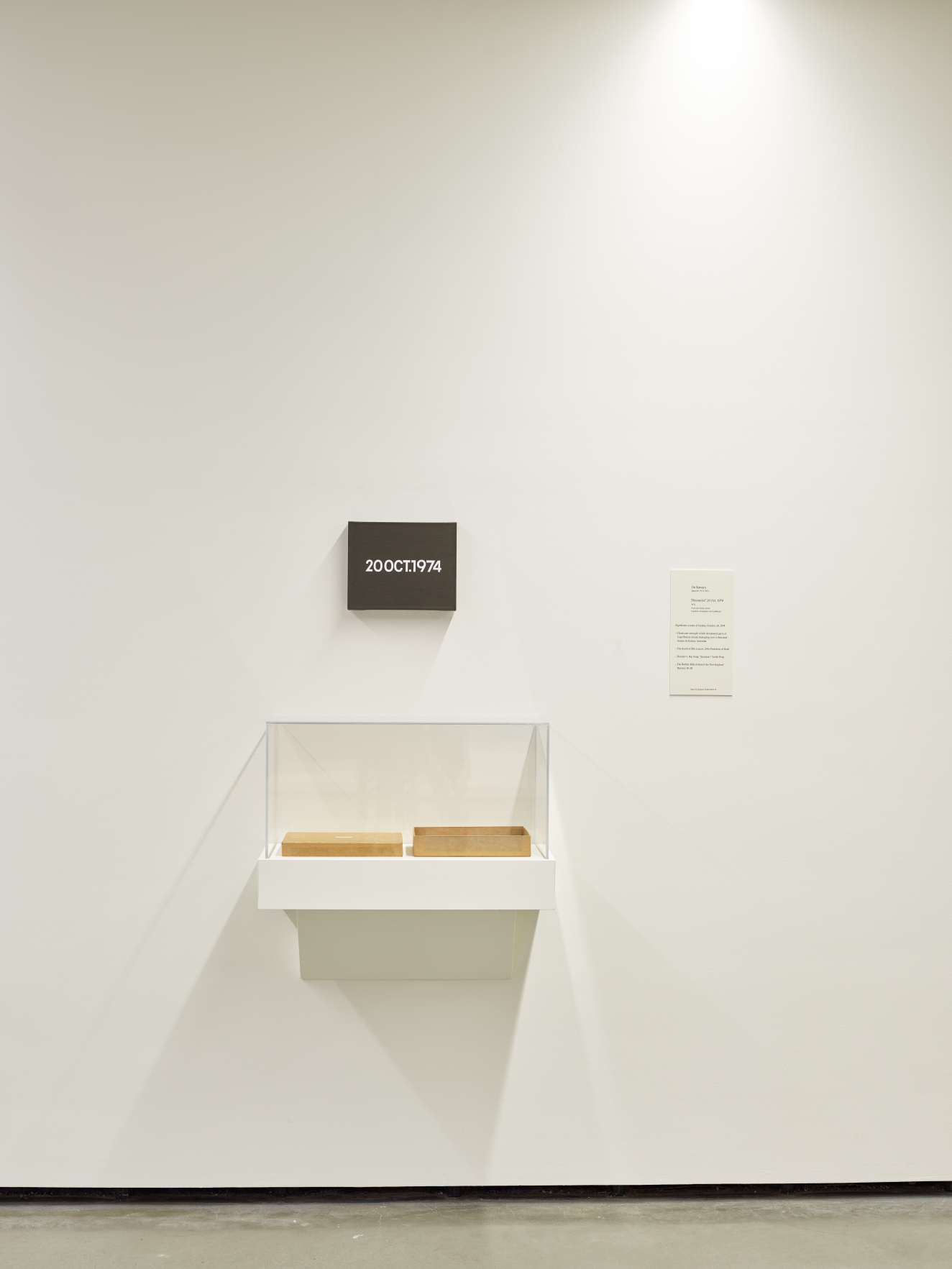  An installation photo depicting part of a white gallery room. A small black panel with illegible text is off-center on the wall, below, an enclosed shelf holds two rectangular objects.