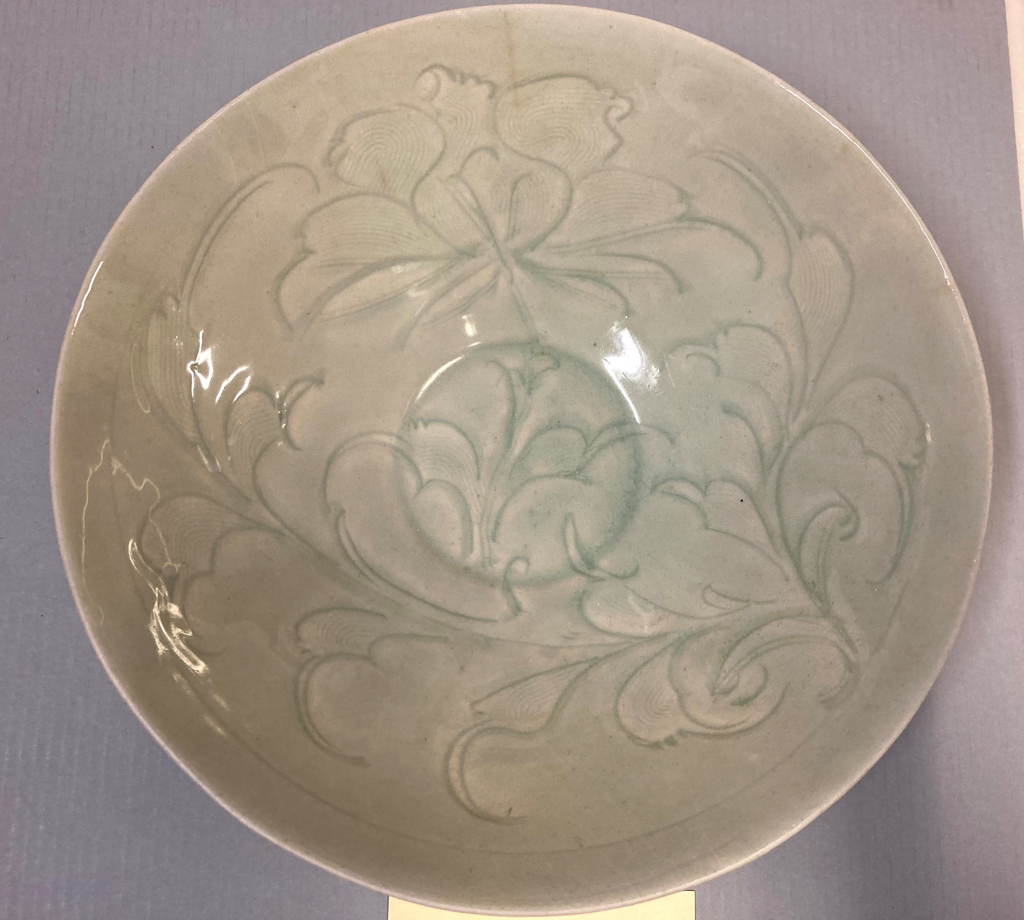 Top view of a round glossy pale gray-green bowl with carved floral motifs which flow into one another. Its base is circular. The plate sits on a purple-gray background.
