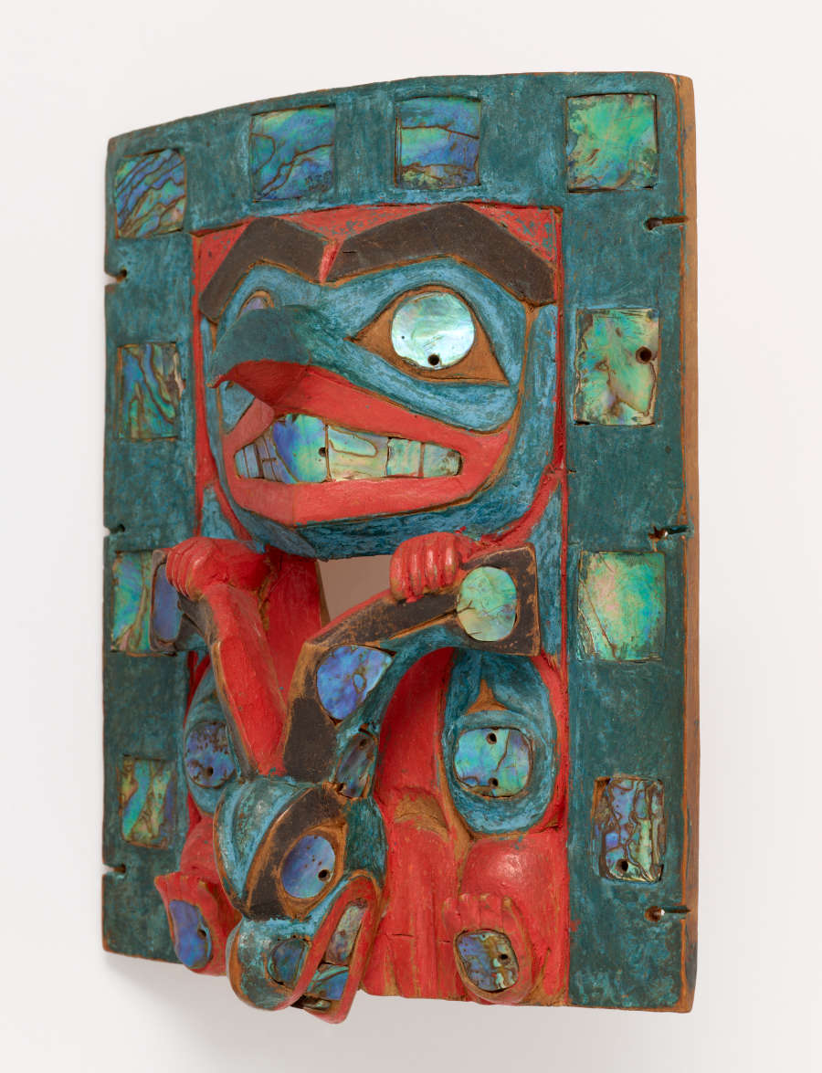 Partial side-view of a painted wooden carving of a face with hands holding onto a deer’s antlers, with a patterned frame made of iridescent shells. Visible is the carving’s three-dimensionality.