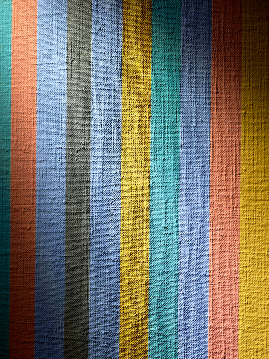 Close-up of painting surface, a spotlight illuminates evenly sized narrow vertical stripes in varied vivid colors, revealing pronounced raised canvas weave and small surface imperfections visible through the matte paint. 