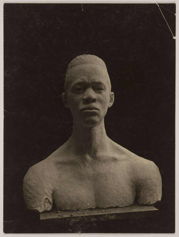 Photograph depicting a chipped bust of a male figure with afrocentric facial features, against a dark background. The face looks contemplative featuring furrowed eyebrows. The body is smooth yet detailed.