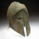 Bronze helmet with a corroded surface which has significant green discoloration. Large sharp eye holes emerge from the narrow nose guard and there is jagged damage by the top.