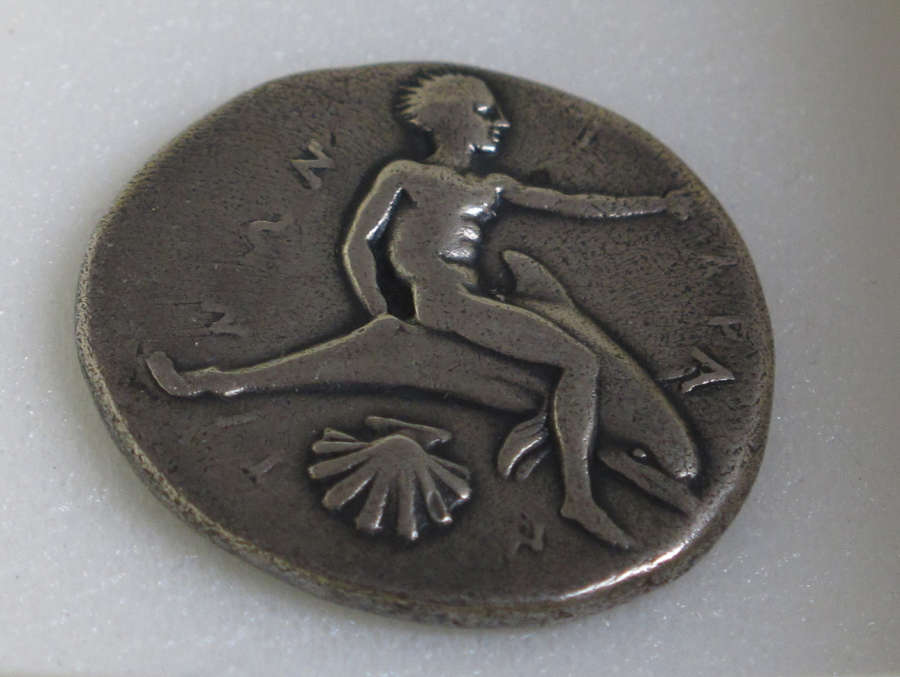 The other side of the silver coin, embossed with an image of a nude man sitting on a dolphin, reaching to the right, above a clamshell. Lettering surrounds his figure.