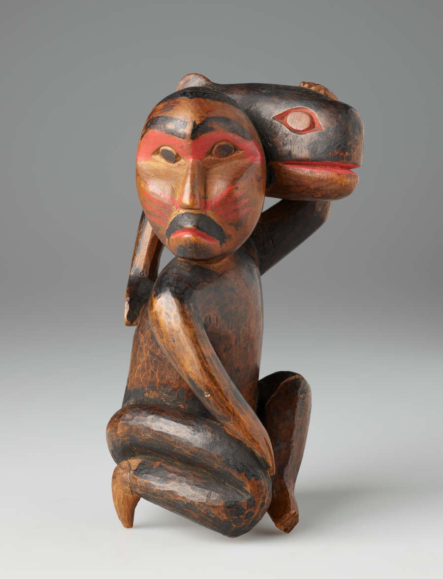 Wooden sculpture of a kneeling bearded man looking aside, holding a small whale against his head. Both man and fish are detailed in partially worn red and black paint.