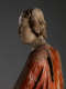 A profile view of a weathered wooden statue of a figure with a solemn expression, with red and green drapery.