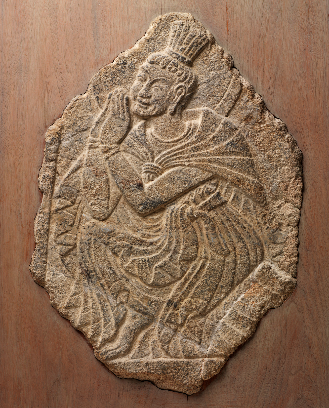 Beige stone relief carving of a kneeling figure with a serene expression wearing flowing robes, with hair bound into a tall bundle and whose hands are clasped in reverence. 