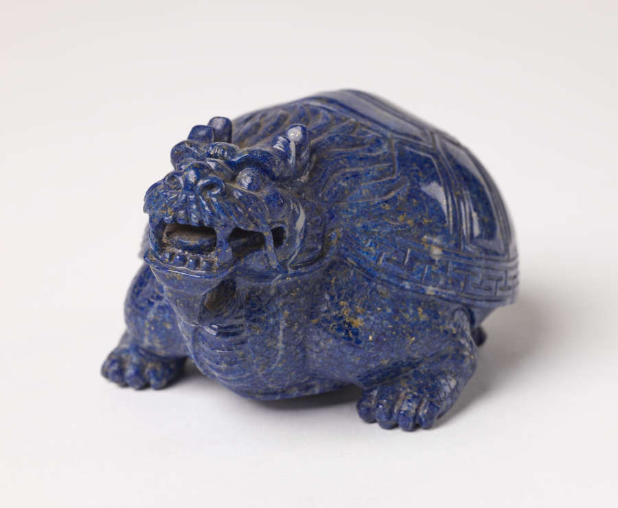 A head-on view of a blue sculpture of a smiling horned tortoise, whose surface is covered with carved geometric patterns.