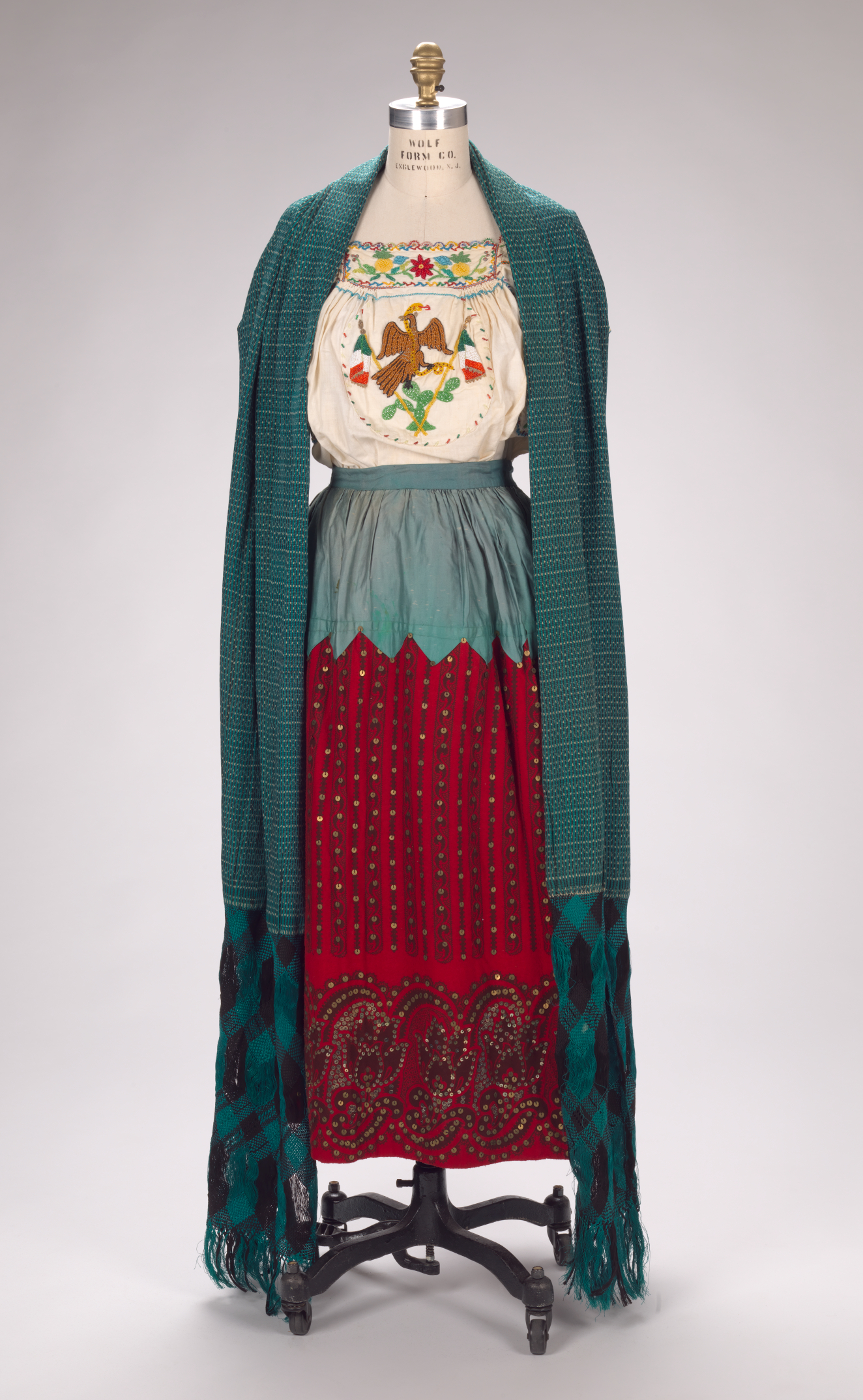 Floor-length, blue shawl layered over a colorful eagle emblem embroidered on a white blouse tucked into a long cinched skirt with zig-zag blue upper half and vibrant red lower half.