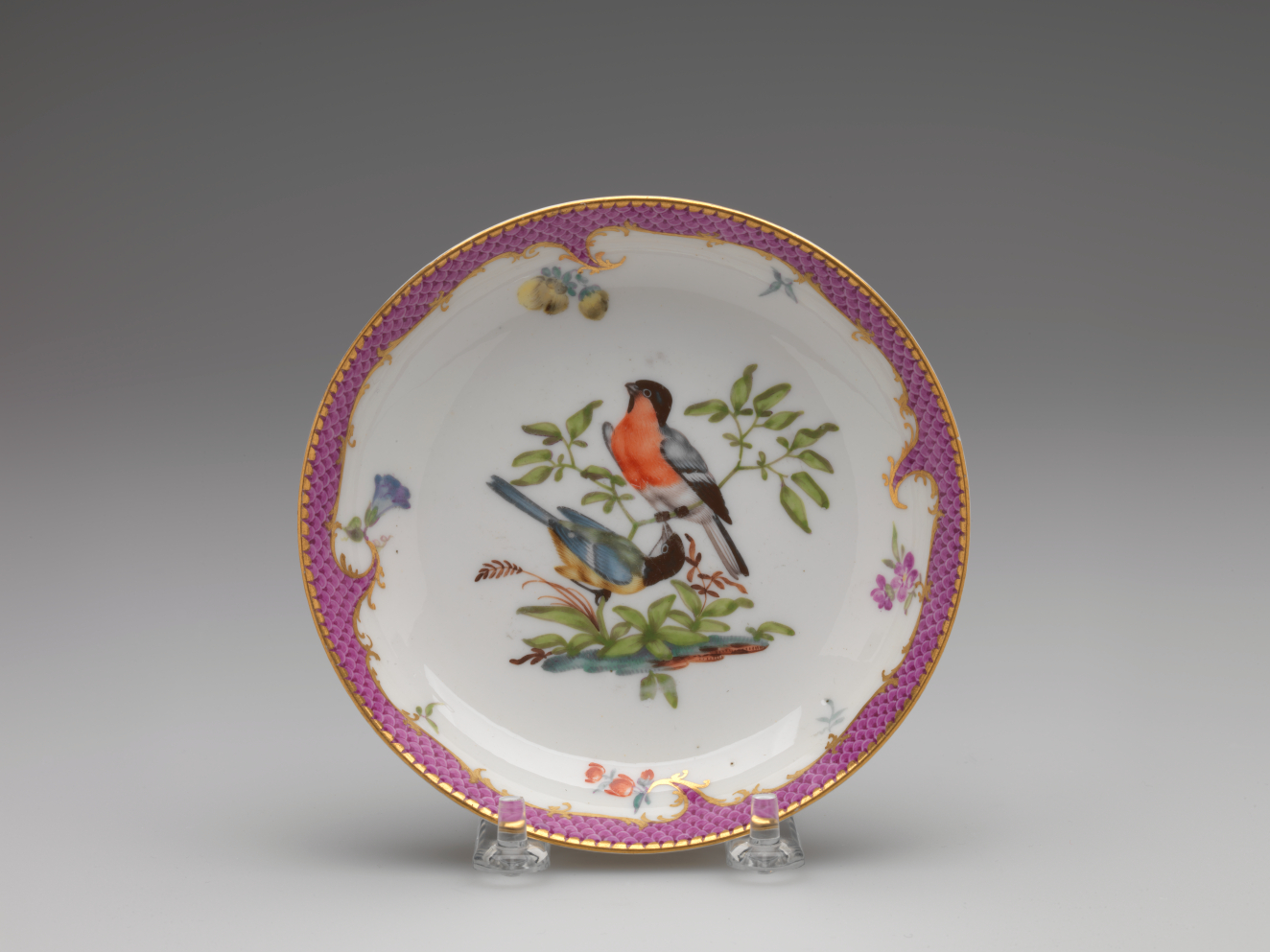 A white plate with a gilded and pink edge and floral patterns. Center design features two birds perched on leafy branches.
