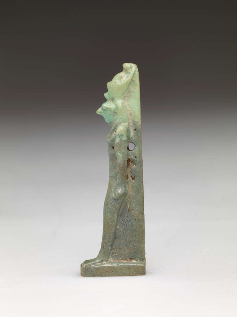 Side-view of a light-jade sculpture of a standing person with a tall head, narrow torso, and straight arms. The left leg has brown specks while the headdress is pale.