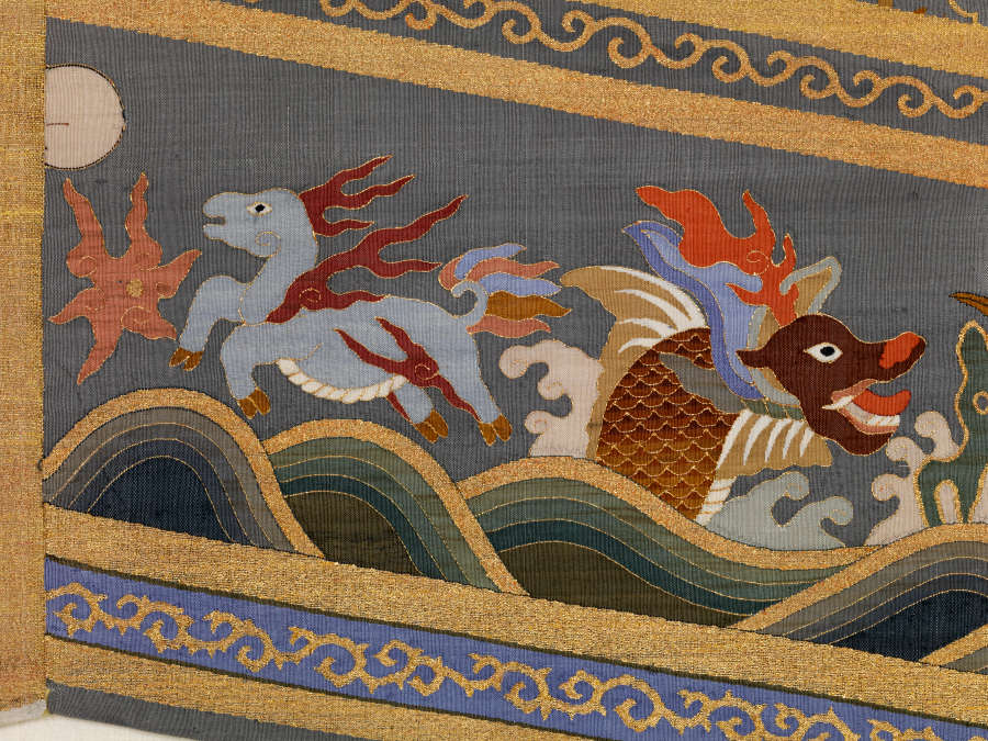 Border detail of the triangular tail of the robes back featuring a red dragon and leaping animal in waves against earthy-pastel clouds encased in golden striped borders.