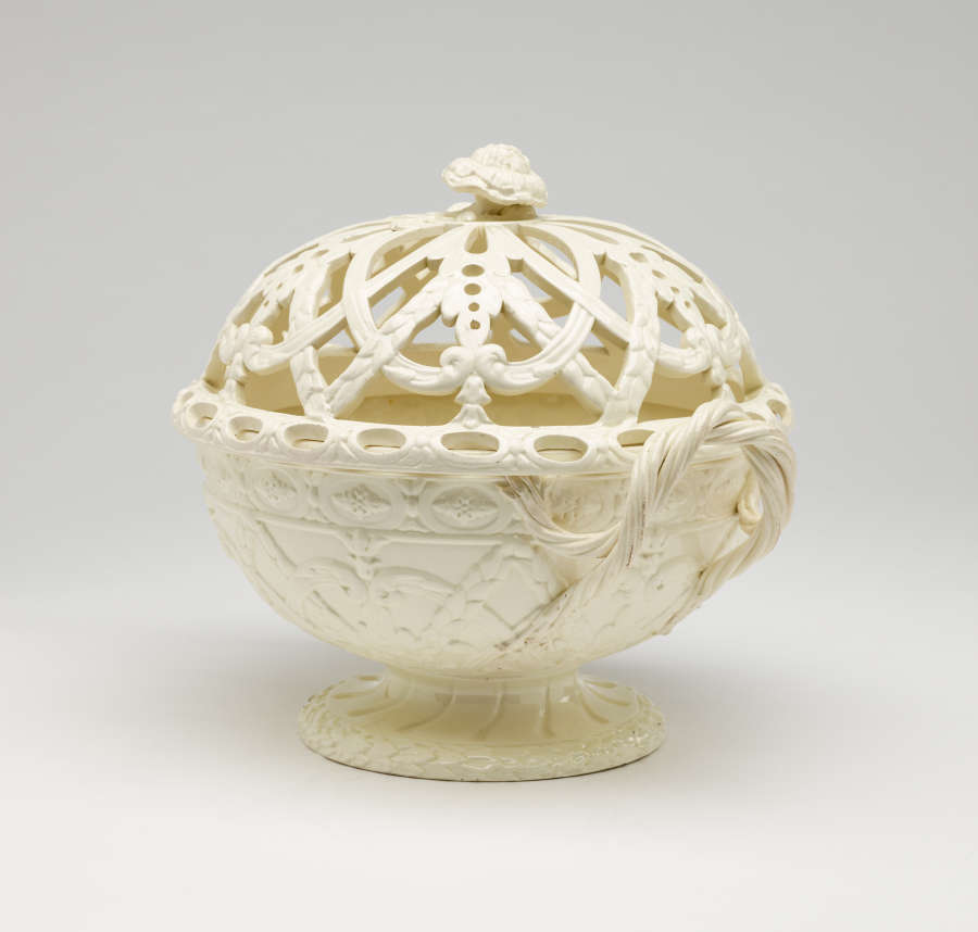 A cream-colored basket with sculptural woven handles, sculpted foot, and a lid with cutout designs.