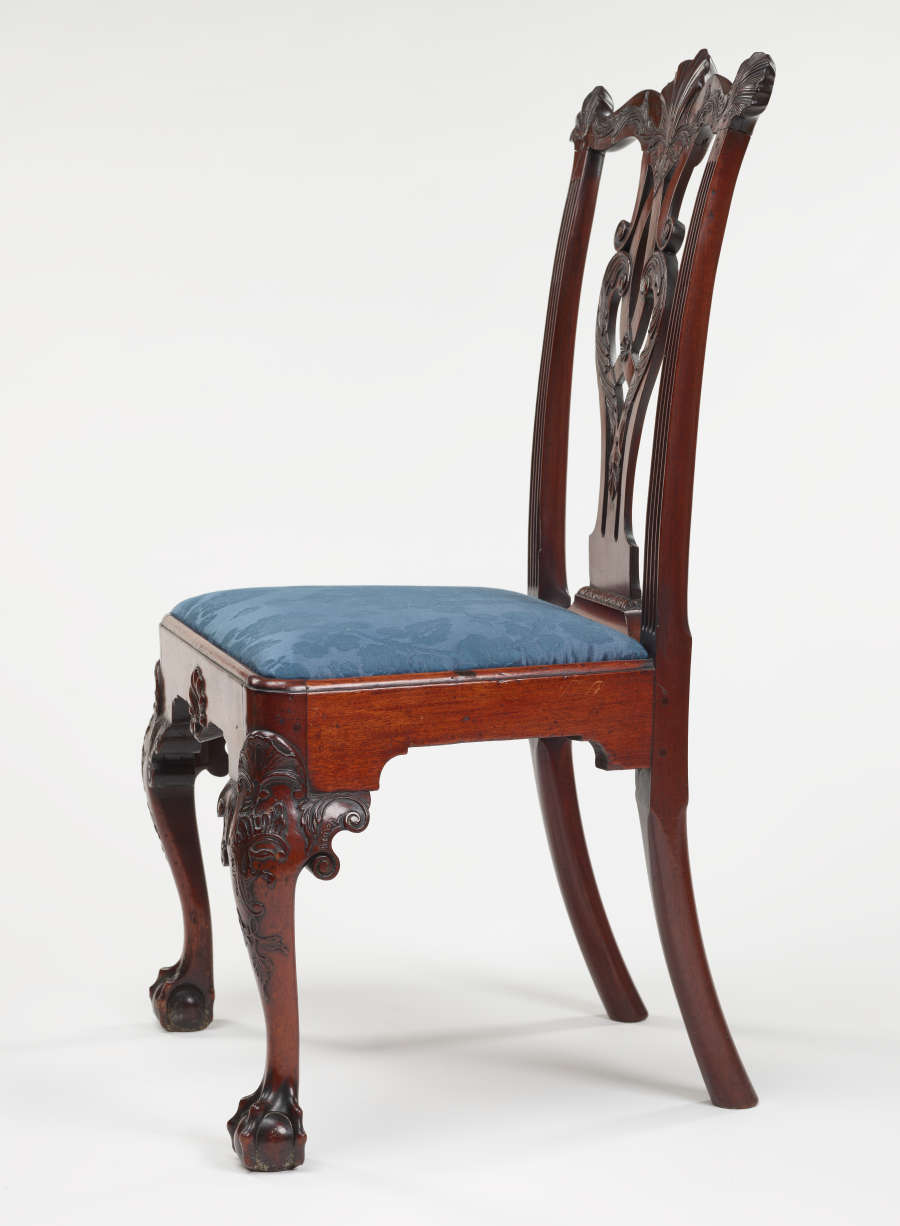 A side view of a wooden chair with an intricately carved open back and a blue upholstered seat, supported by clawed feet.