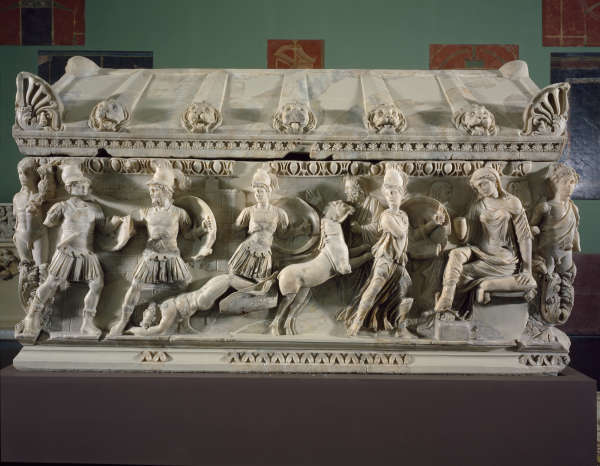 A marble coffin with an angled roof and deep-set carvings depicting human and animal figures engaged in battle. 