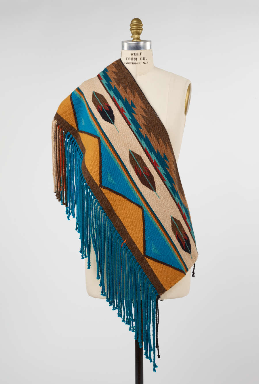 Multicolored woven tapestry with patterning and fringe, draped diagonally across the torso of a mannequin. The mannequin's right shoulder is bare with fringed ends cascading off the mannequin’s left-side. 