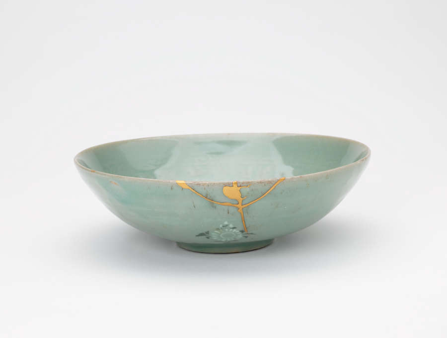 Side-view of a tilted, light-teal, stout glazed bowl, with green-blue floral motifs, faded floral patterns, and gold patched cracks along a top-edge.