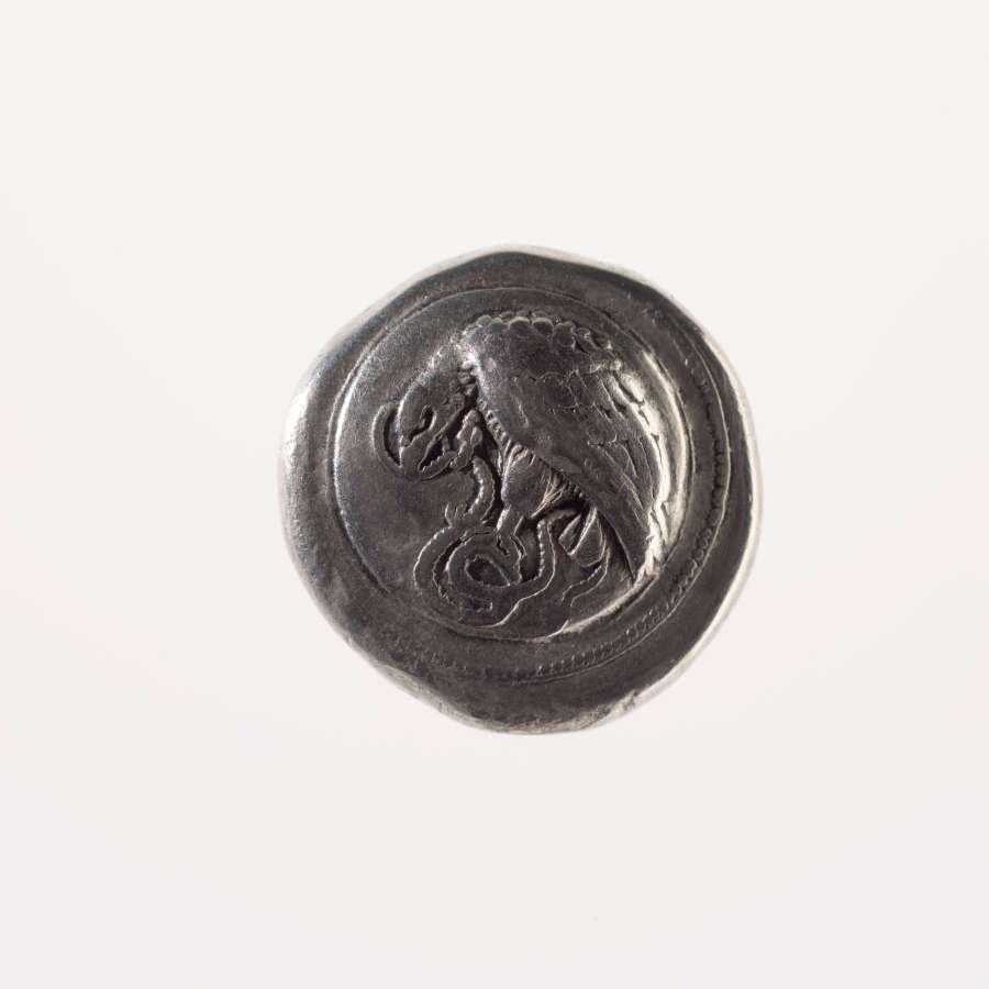Irregular round silver coin embossed with a depiction of an eagle, curved against a circular depressed border, with a snake in its hands and mouth.
