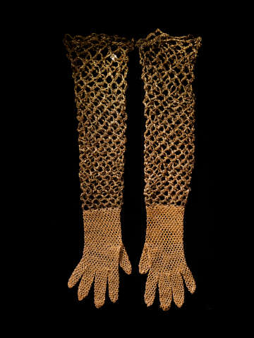 A pair of long gold mesh gloves on a dark surface, with fine chain-linked netting from the fitted hands extending to the open-linked upper arms.