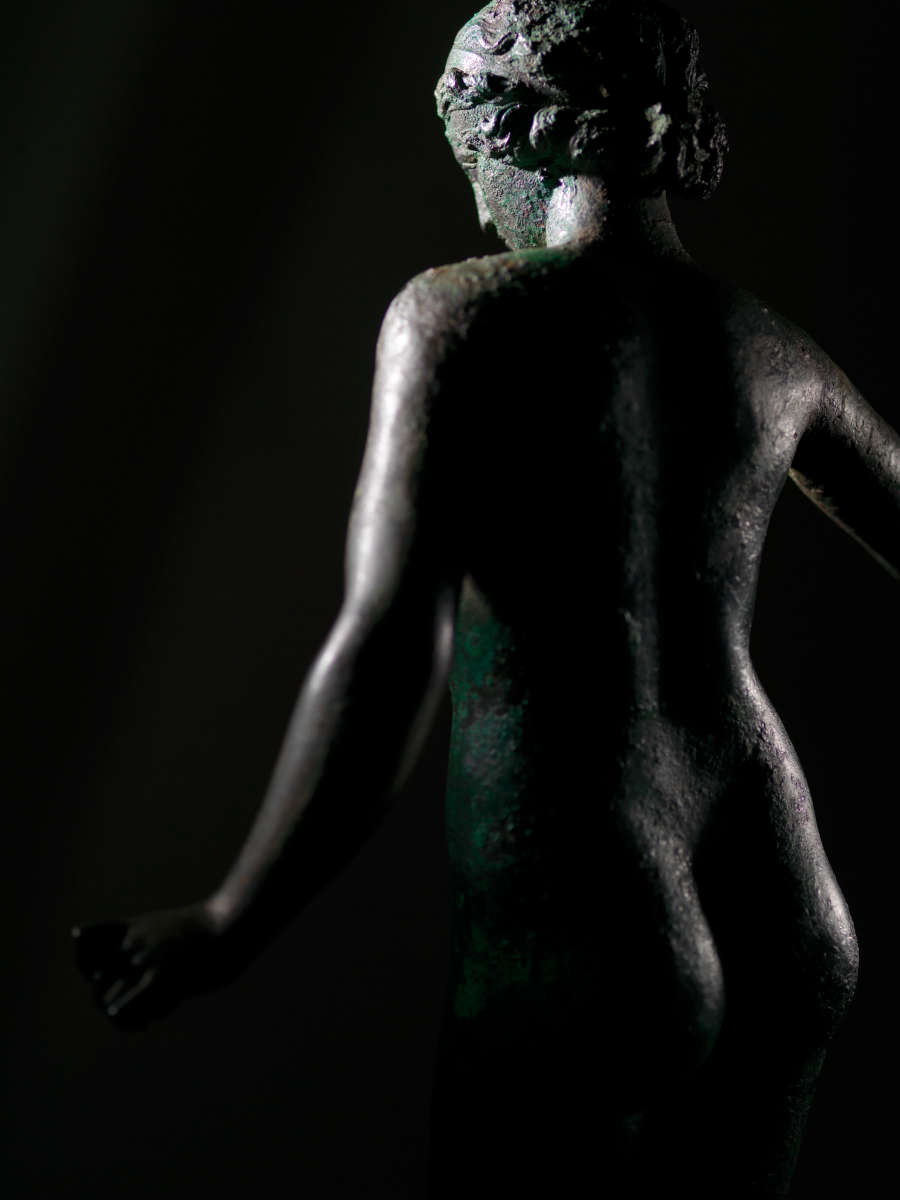 Back view of a tarnished bronze statue of a standing nude woman, her arms away from her body, as she emerges from the darkness. The lighting amplifies the texture of the statue’s surface.