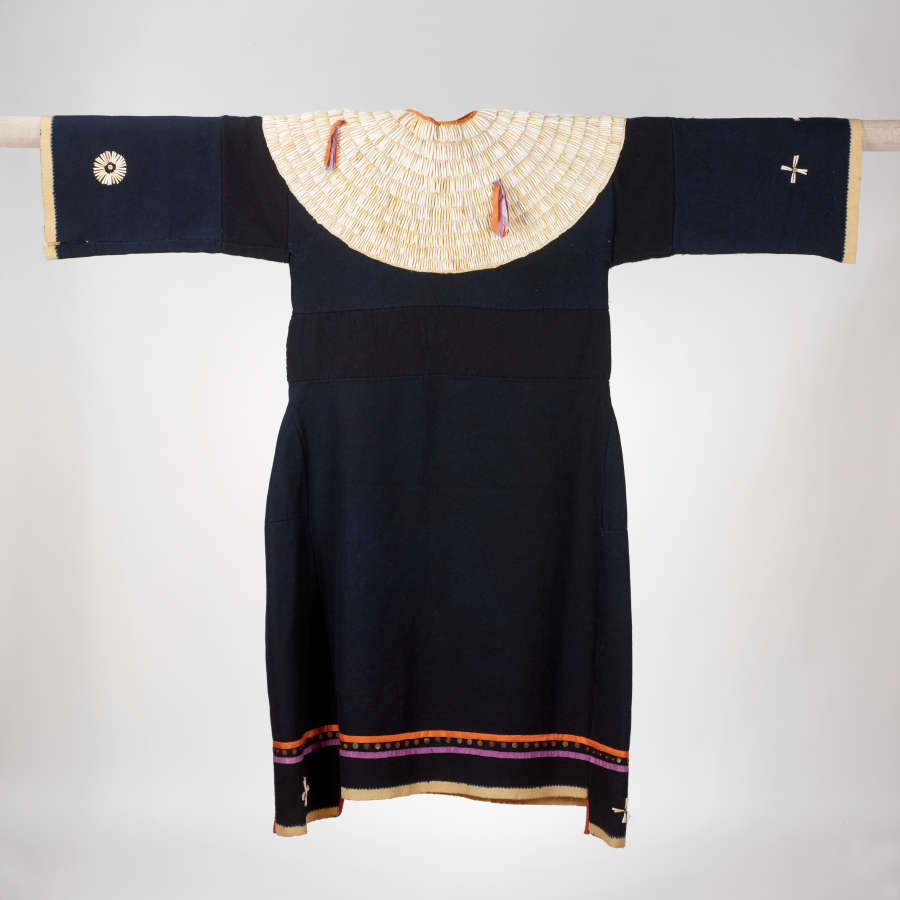 Black, long dress hung on a rod. The collar consists of several off-white concentric rows with two tassels, the sleeve ends feature geometric motifs, and the hem features multicolored stripes.