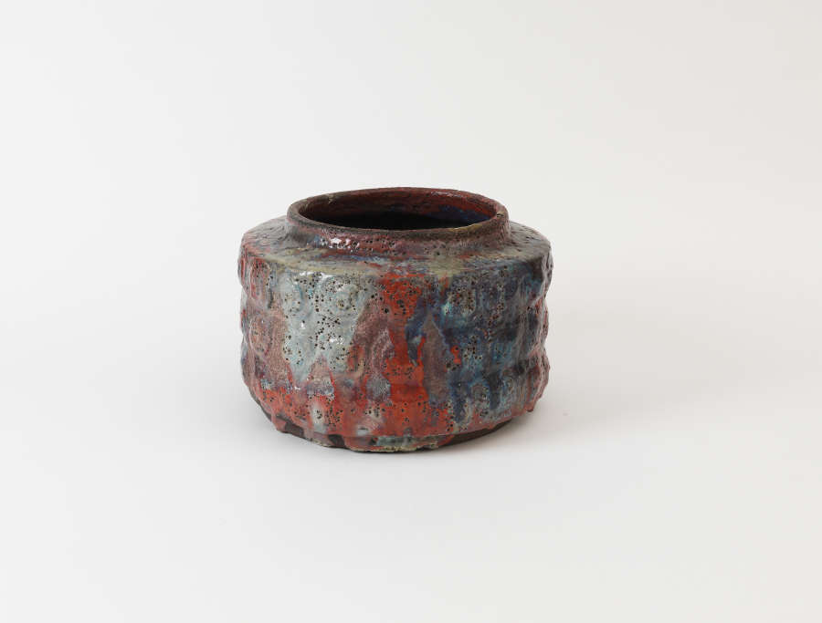 Small, cylindrical ceramic jar glazed with a glossy finish with a rough, ribbed surface covered in pores, the earthy red clay textured with patches of blue and gray tones.