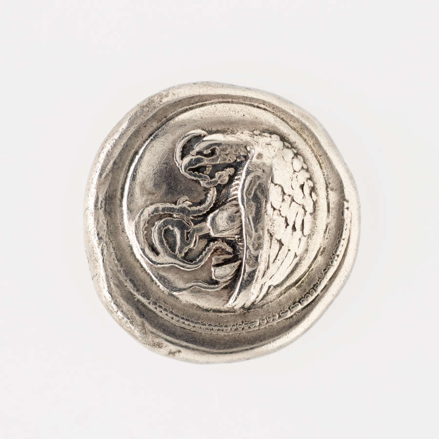 Irregular round silver coin embossed with a depiction of an eagle, curved against a circular depressed border, with a snake in its hands and mouth.