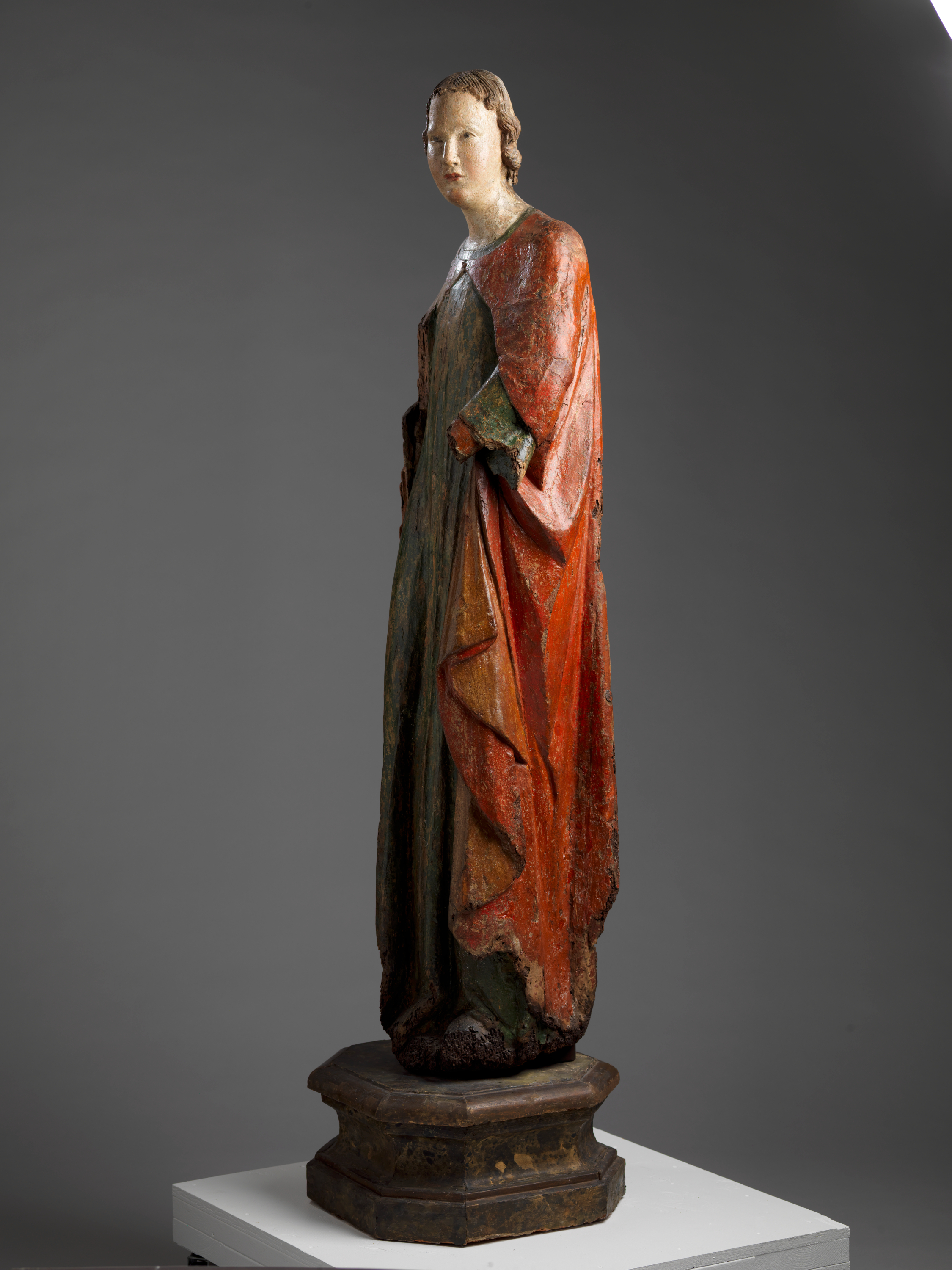 A tall, weathered wooden statue of a robed figure with a serene expression, draped in green and red garments, standing upon an octagonal base.
