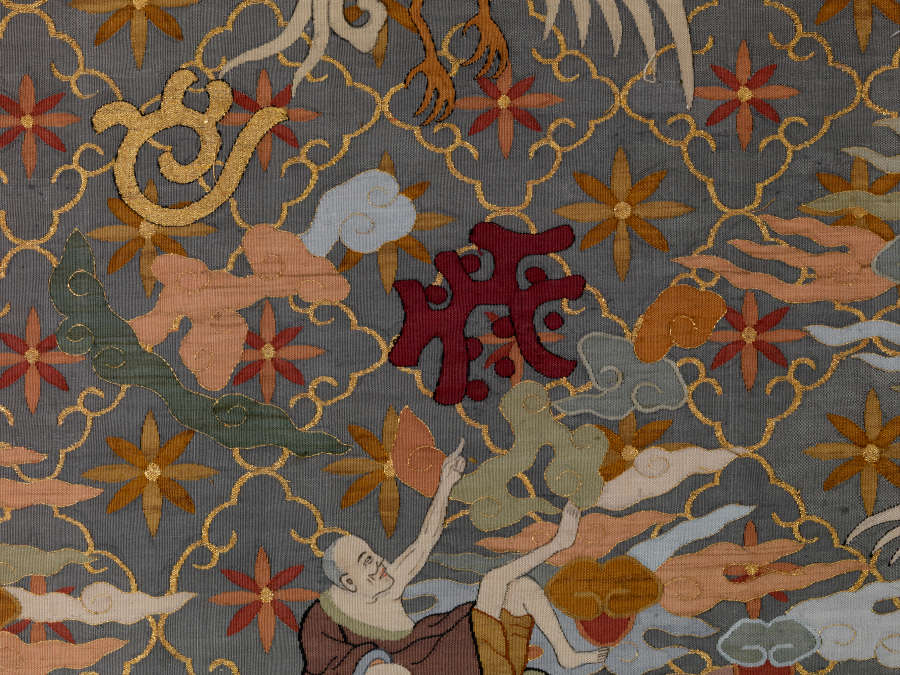 Detail of the blue-gray robe’s center, featuring a reclining robed monk amongst wispy clouds pointing upwards towards abstract earthy-pastel symbols, against thin diagonal grid pattern with earthy floral motifs.