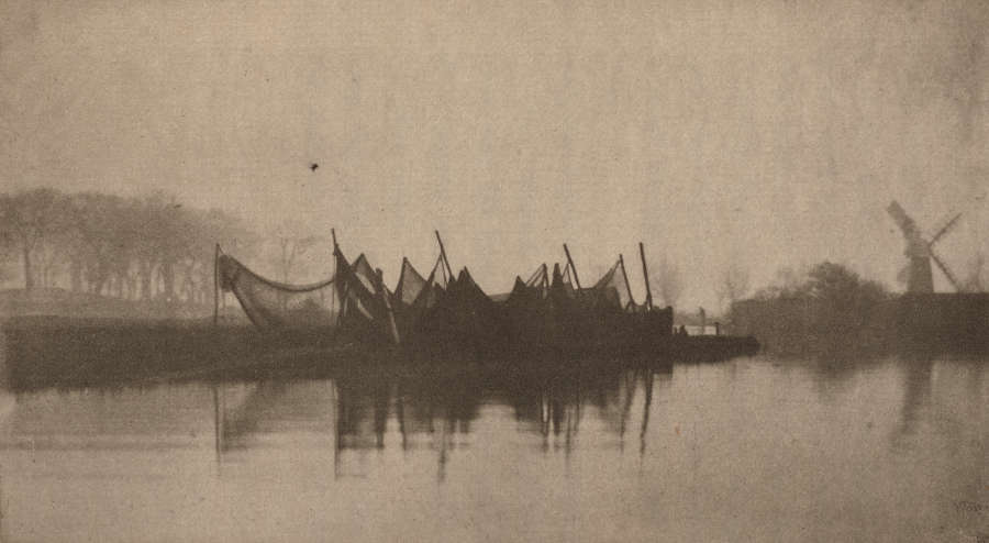 A misty riverside with a structure made of nets stretched across poles at the shore. In the distance, there is a windmill to the right and trees to the left.