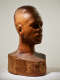 Three quarter view of a textured, but glossy, wooden-carved bust depicting a male figure with afrocentric facial features. The expression is cast in shadow, highlighting the side of the head.