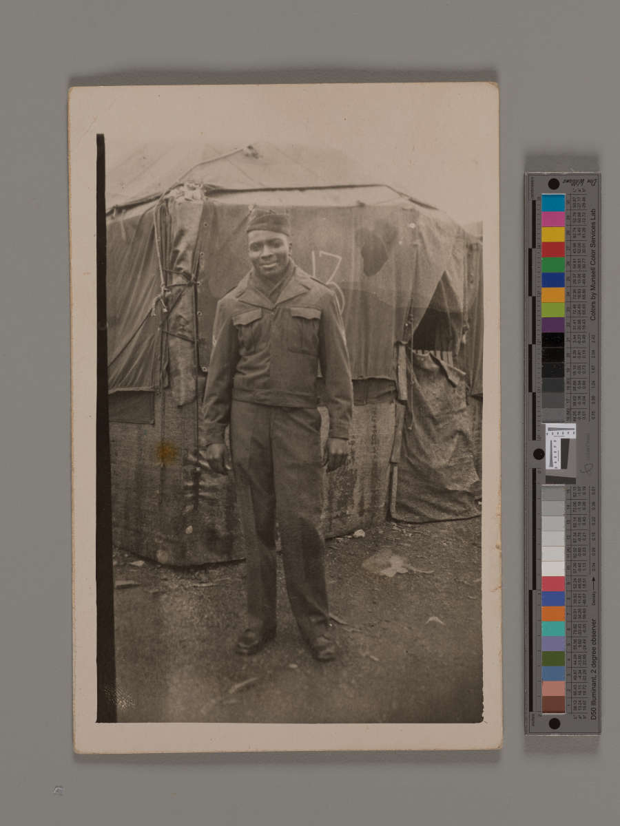  A handsome young Black soldier in uniform stands smiling in front of a military tent.
