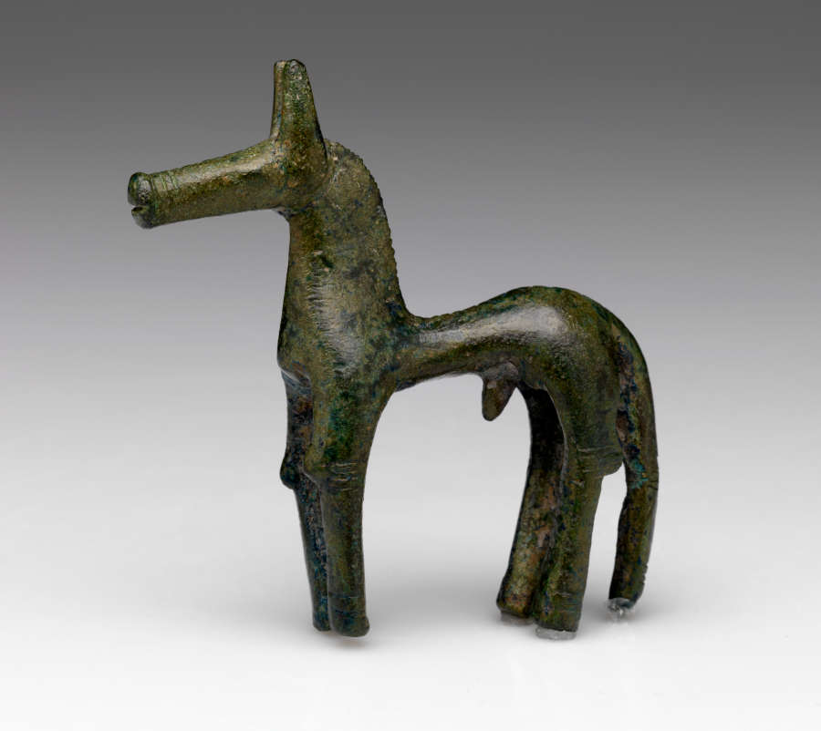 Side-view of a bronze horse figurine with a green patina, and an exaggeratedly thin torso, thick neck, long legs and tail and long narrow snout.